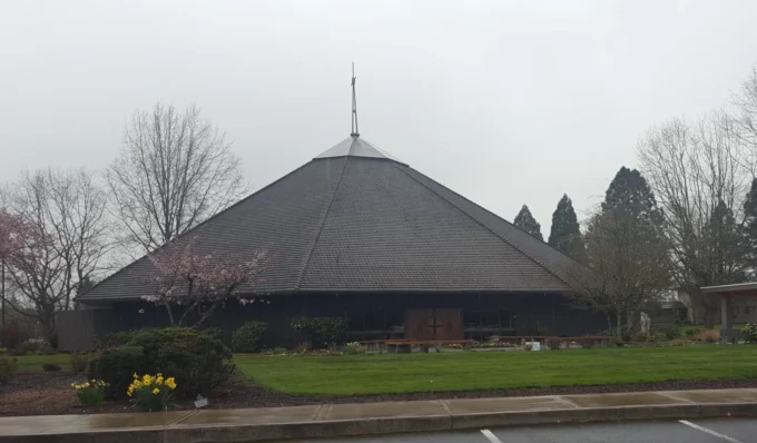 A church with a steep pyramid-shaped shingled roof, wooden cross above entrance, surrounded by lawn and spring flowers