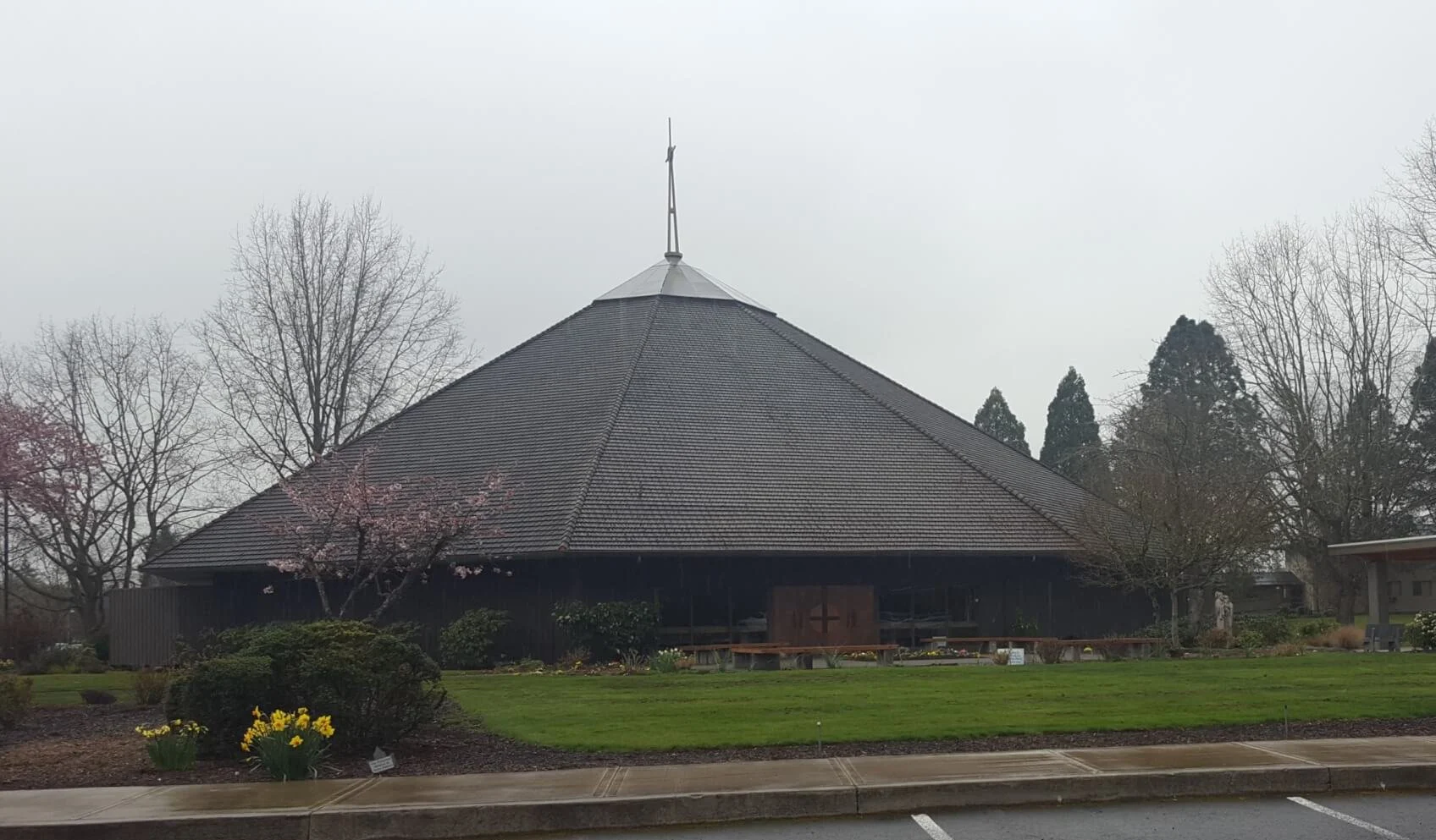 A church with a steep pyramid-shaped shingled roof, wooden cross above entrance, surrounded by lawn and spring flowers