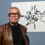 Man with glasses and brown leather jacket stands before an abstract black ink drawing on a gallery wall