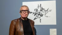 Man with glasses and brown leather jacket stands before an abstract black ink drawing on a gallery wall