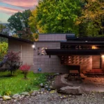 Single-story home with a low, angled overhanging roof and patterned concrete block walls, surrounded by autumn trees