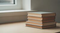 Stack of five hardback books with plain cloth covers on a pale timber surface by a window