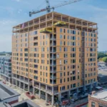 Tall wood-framed high-rise under construction with a crane on top, surrounded by lower city buildings