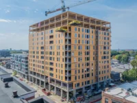 Tall wood-framed high-rise under construction with a crane on top, surrounded by lower city buildings