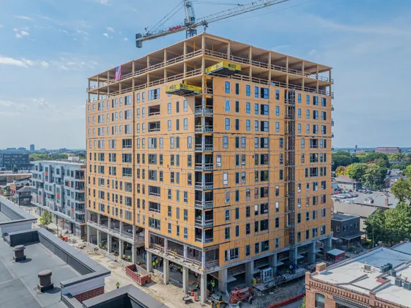 Tall wood-framed high-rise under construction with a crane on top, surrounded by lower city buildings