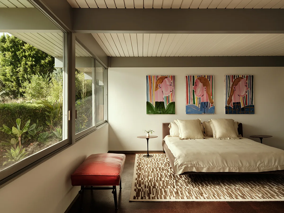 Bedroom with a wall of glass, a low bed and a triptych of colourful portrait paintings above the headboard