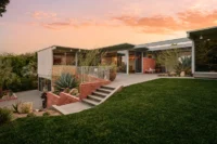 Mid-century modern Adelman Residence at dusk with glass walls, a lawn and brick planters under a pink sky
