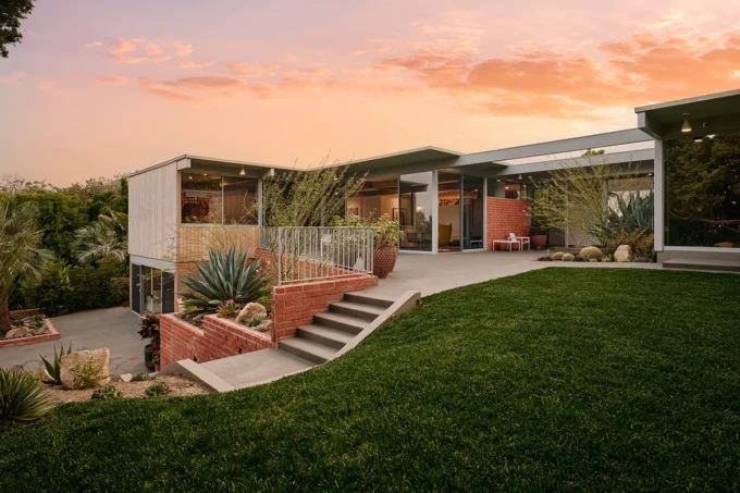 Mid-century modern Adelman Residence at dusk with glass walls, a lawn and brick planters under a pink sky