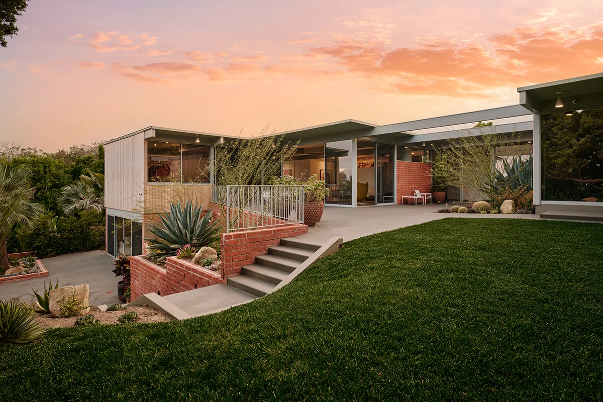 Mid-century modern Adelman Residence at dusk with glass walls, a lawn and brick planters under a pink sky