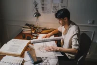 Young architect at a lamp-lit desk sketching over large drawings with books and rolled plans around him