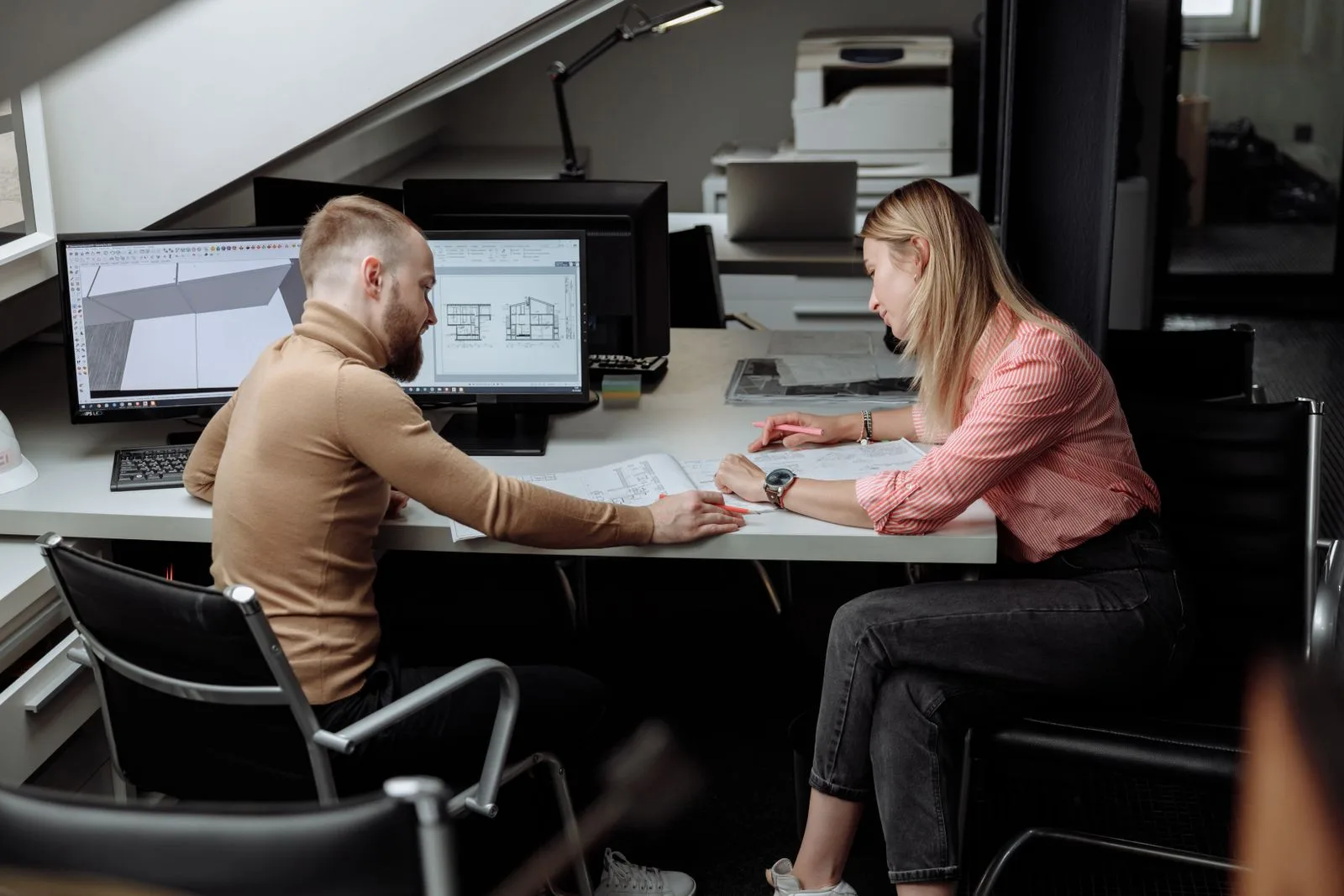Two architects sitting at a desk with monitors and printed plans, discussing a drawing