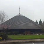 A church with a steep pyramid-shaped shingled roof, wooden cross above entrance, surrounded by lawn and spring flowers