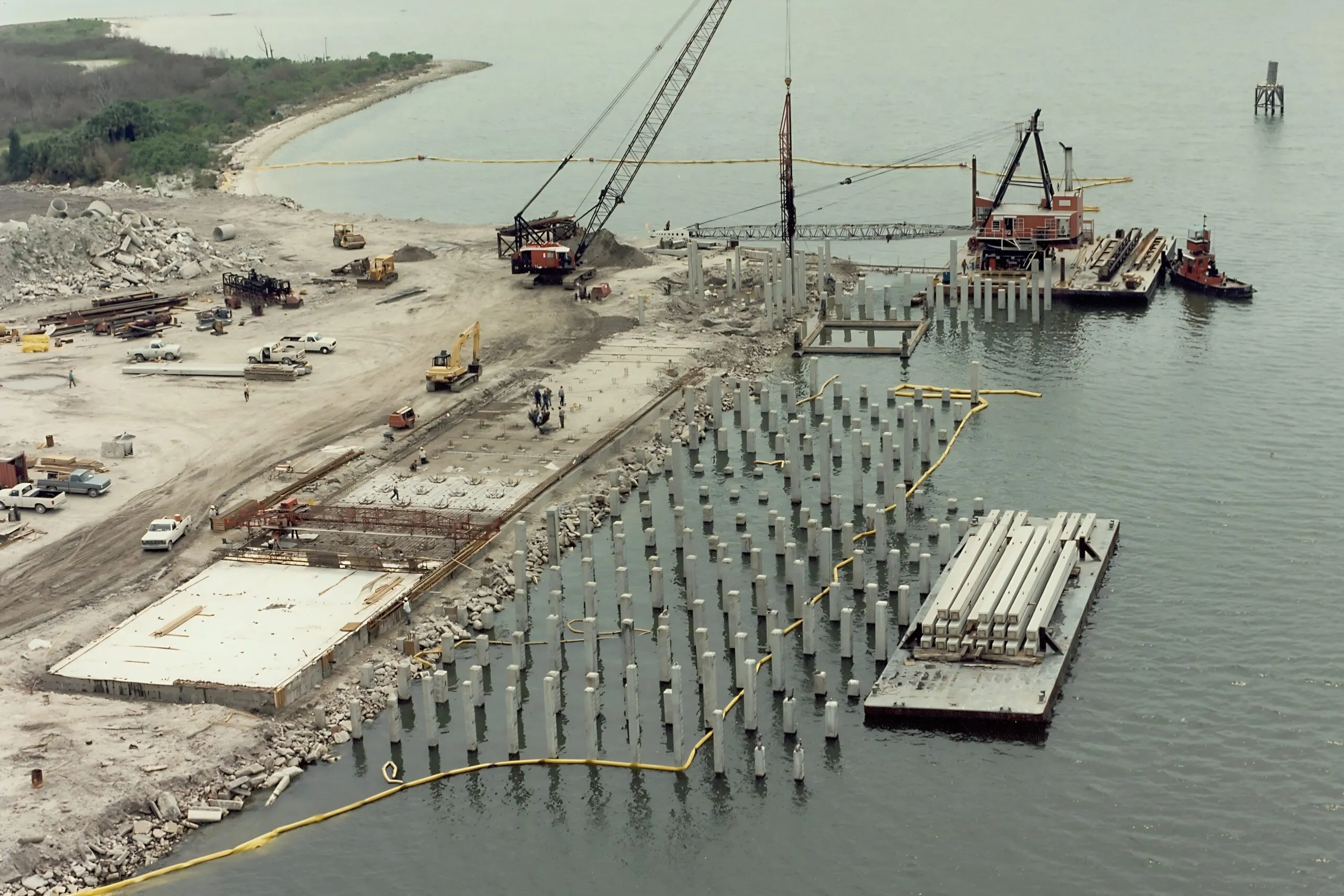 Overhead shot of a shoreline building site showing pile grids, cranes, trucks and a floating barge with pipes
