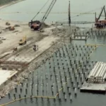 Aerial view of a waterfront construction site with rows of concrete piles driven into the seabed and cranes on a barge