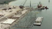 Aerial view of a waterfront construction site with rows of concrete piles driven into the seabed and cranes on a barge