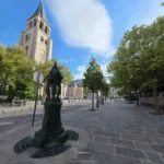 Cobblestone plaza with an ornate fountain pillar, trees, and a stone church tower under blue sky