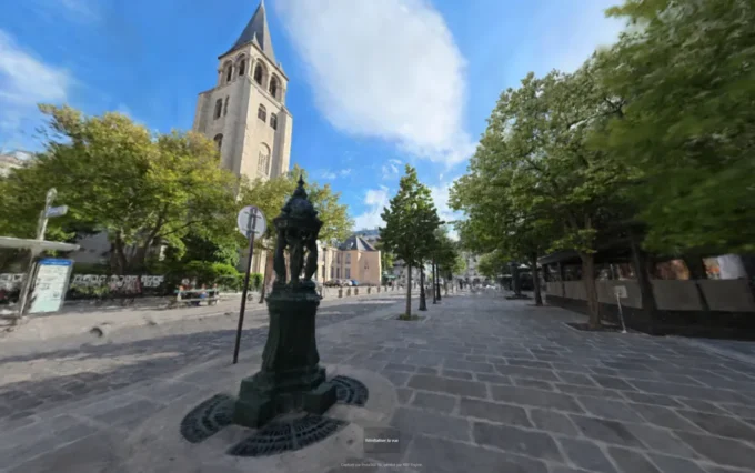 Cobblestone plaza with an ornate fountain pillar, trees, and a stone church tower under blue sky