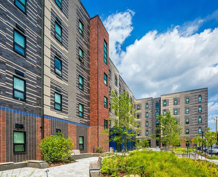 Corner view of a brick apartment building with landscaped courtyard, benches, and shrubs under a partly cloudy sky