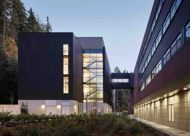 Dusk view of a dark-clad building with a glass stairwell connected by an elevated walkway to a brick building