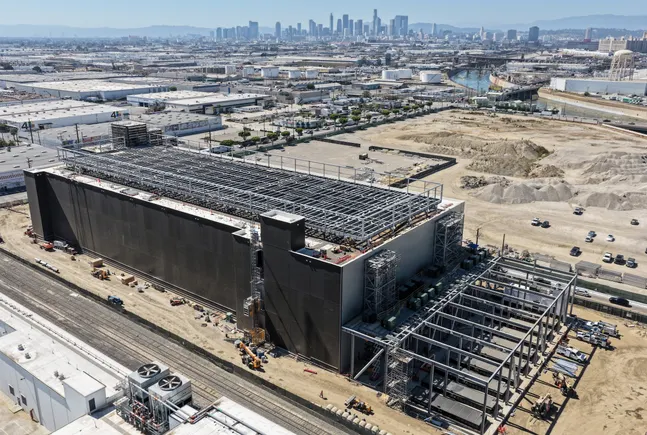 Aerial view of a large industrial building under construction with steel framing, near a city skyline