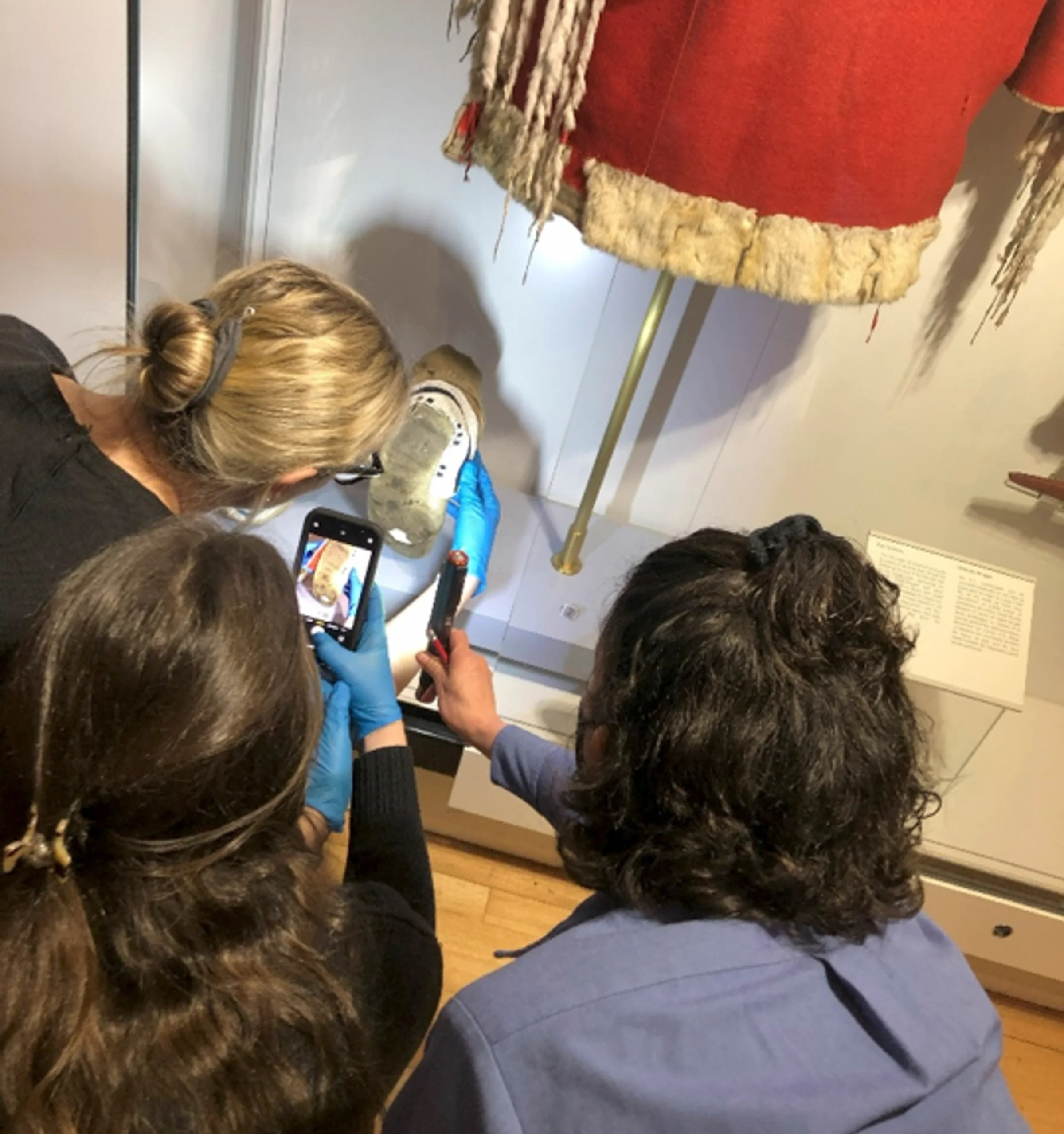 Three people in gloves examine a beaded moccasin and fringed garment displayed in a museum case