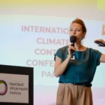 Woman speaking at a podium with a microphone, gesturing beside a climate conference slide
