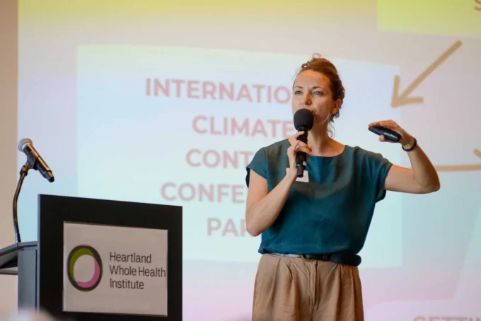 Woman speaking at a podium with a microphone, gesturing beside a climate conference slide