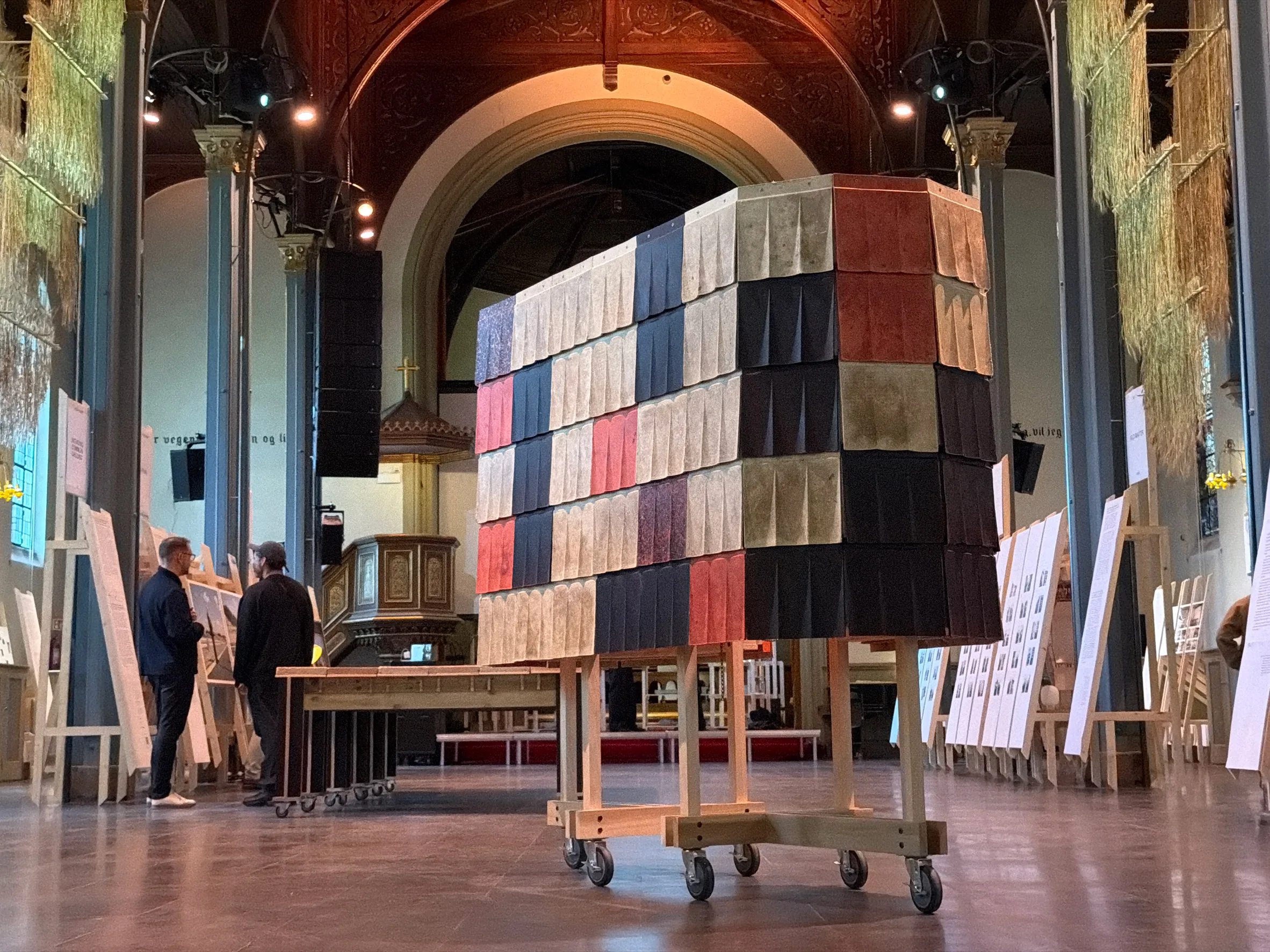 A large cube built from colorful fabric panels sits on a wheeled cart inside a vaulted hall