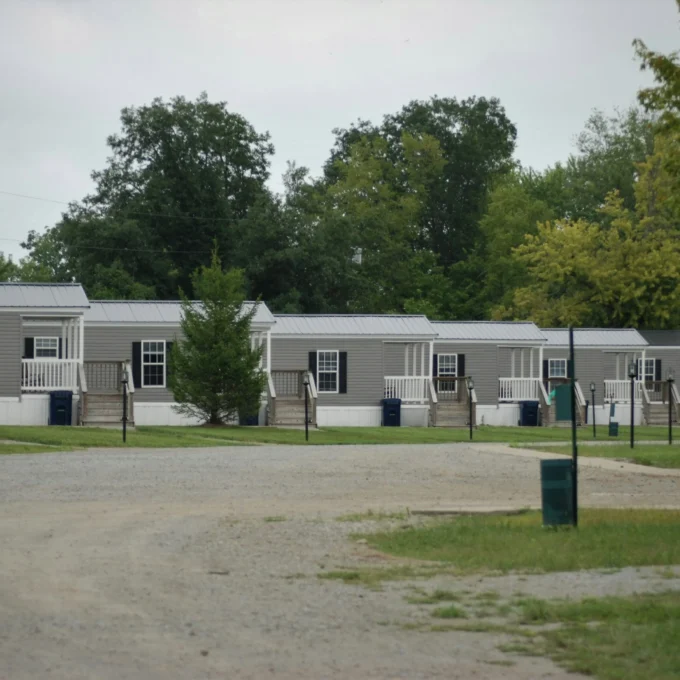 Row of identical gray manufactured homes with white porches along a gravel road