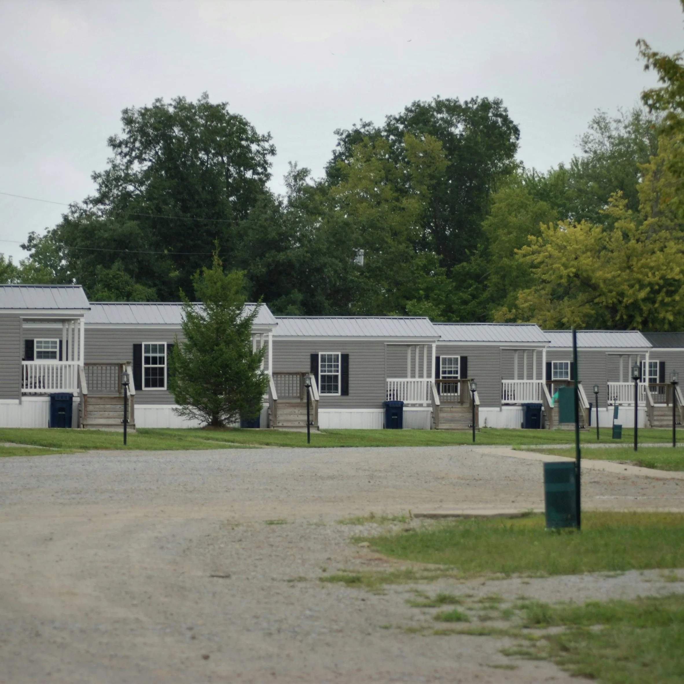 Row of identical gray manufactured homes with white porches along a gravel road