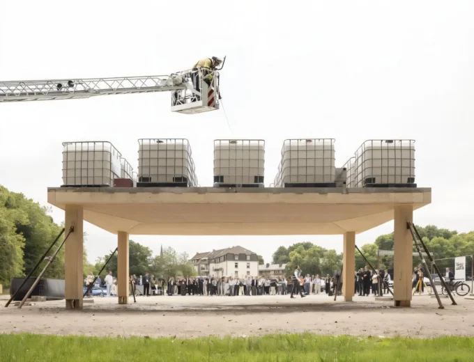 A crowd watches a flat concrete-topped platform on wooden columns, loaded with stacked water tanks, as a crane worker