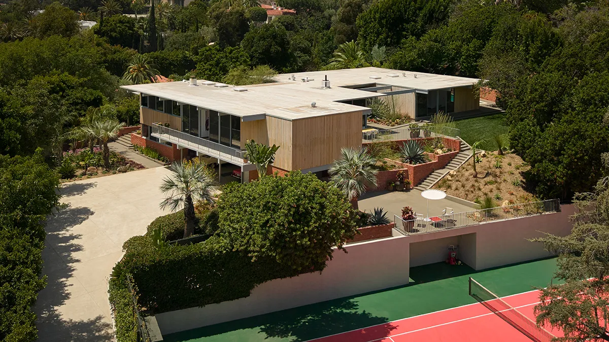 Aerial view of a flat-roofed midcentury home surrounded by desert landscaping, palm trees and a tennis court