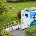A solar-paneled public restroom pod with ramp sits on a path beside a lawn where a person walks a dog