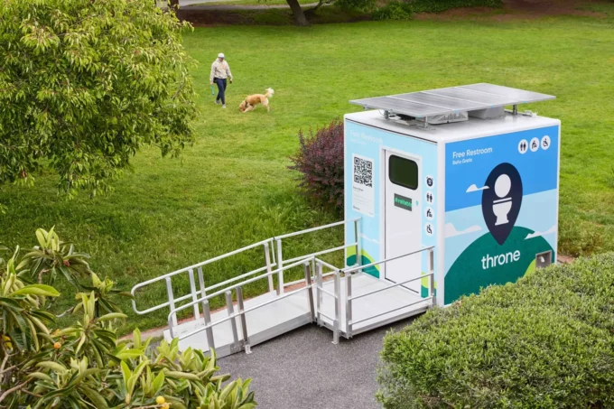 A solar-paneled public restroom pod with ramp sits on a path beside a lawn where a person walks a dog