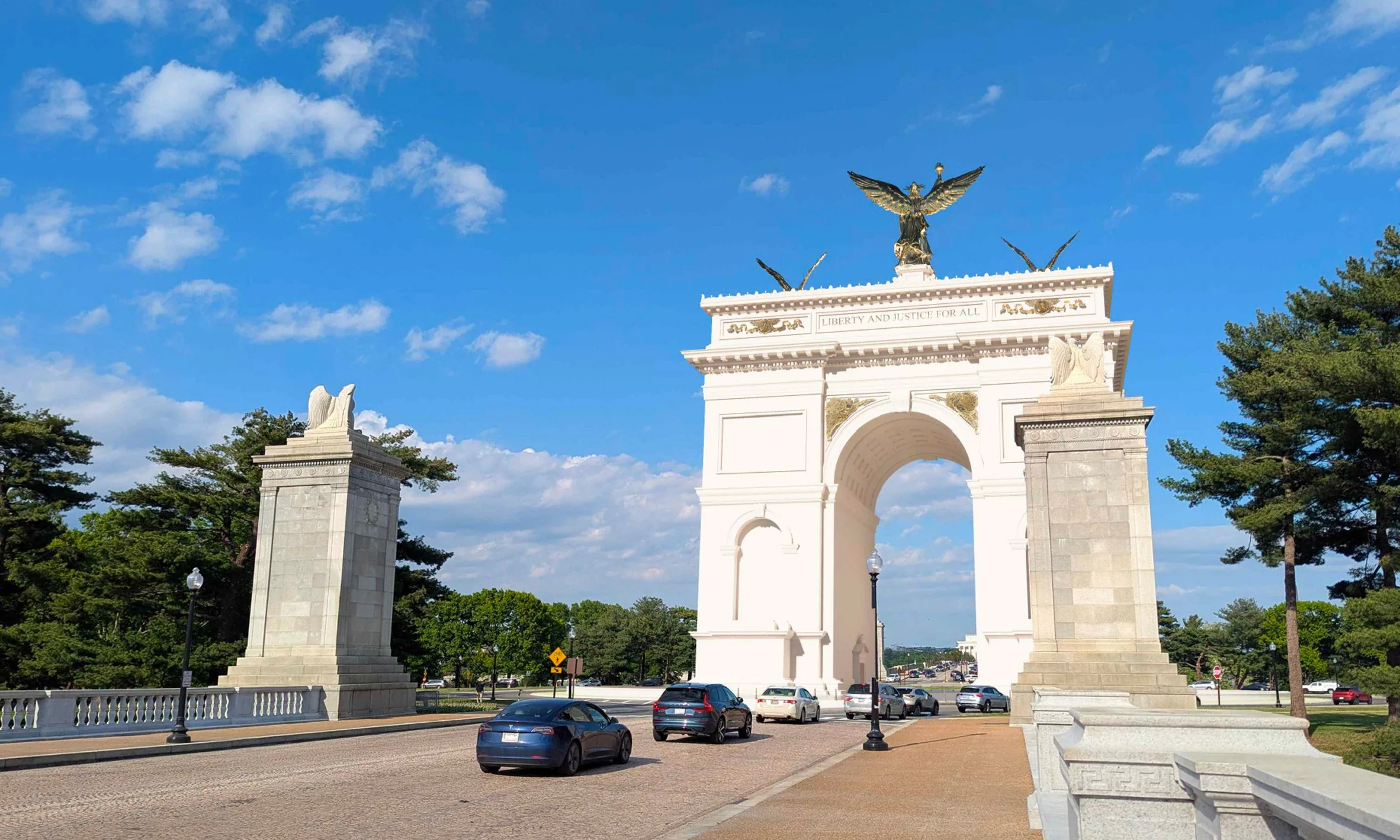White stone arch with a bronze eagle on top, flanked by stone pillars, cars passing beneath on a road
