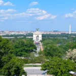 Aerial view over a cemetery and treetops toward a distant white arch, Washington Monument and Capitol