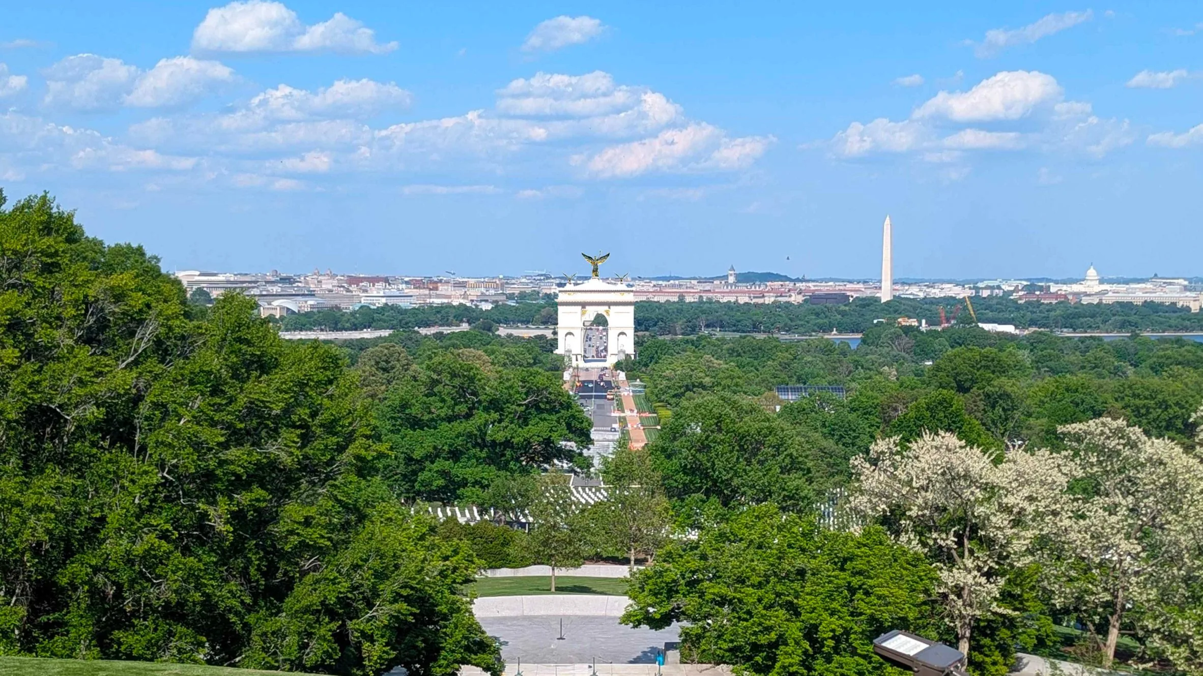 Aerial view over a cemetery and treetops toward a distant white arch, Washington Monument and Capitol