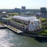 Aerial view of a waterfront building with a rippled glass facade beside a riverside park and city skyline