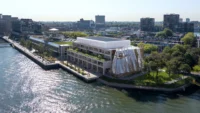 Aerial view of a waterfront building with a rippled glass facade beside a riverside park and city skyline