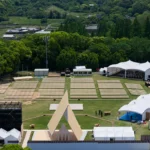 Aerial view of a triangular timber pavilion at a festival site surrounded by tents, lawns and forested hills