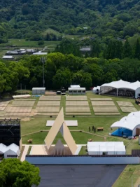 Aerial view of a triangular timber pavilion at a festival site surrounded by tents, lawns and forested hills