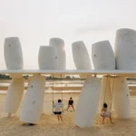 White perforated cylinders above and below a thin roof plane with two swings on a sandy beach at dusk, Seaside Swing Installation by XBTW OFFICE