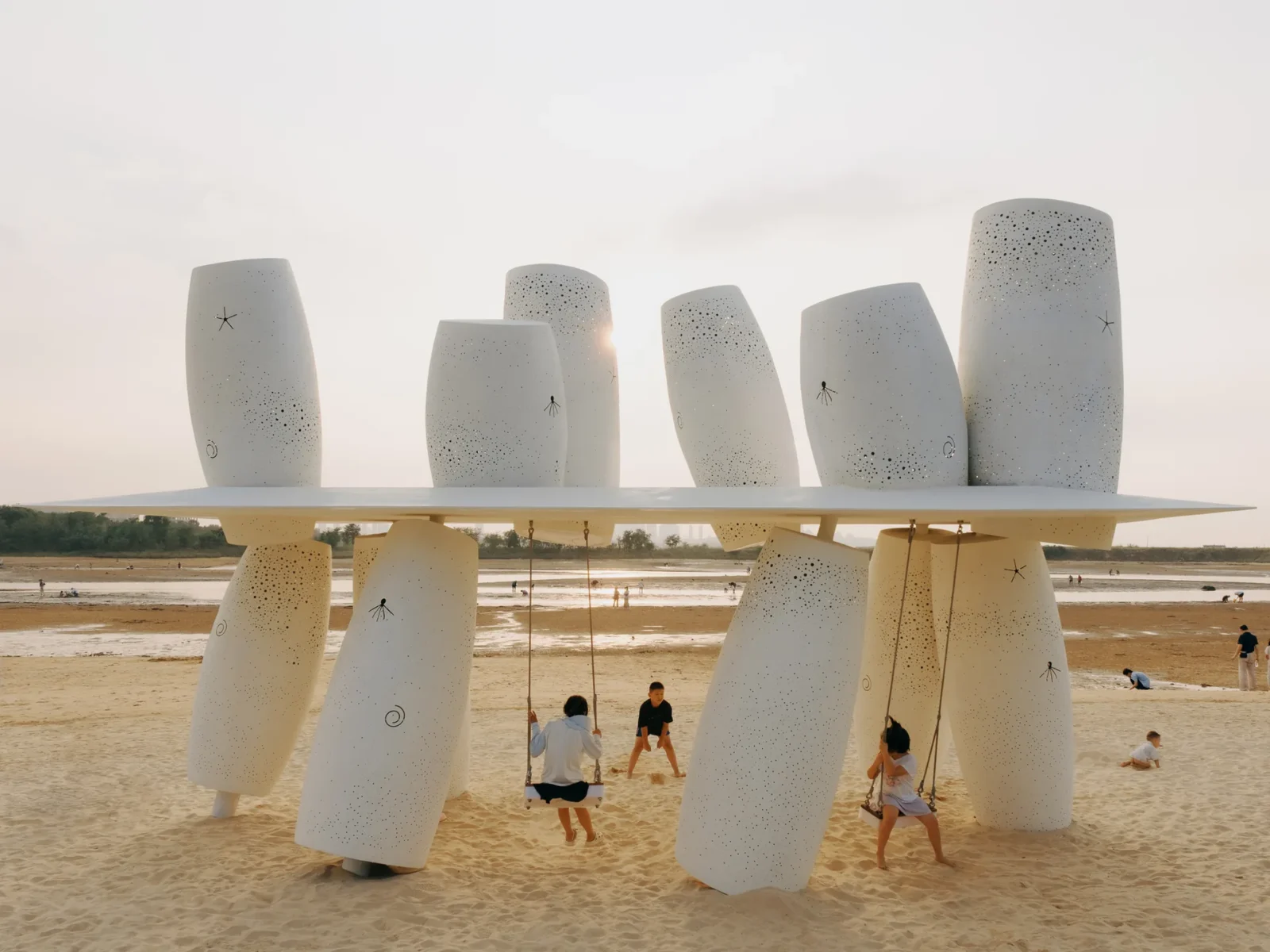 White perforated cylinders above and below a thin roof plane with two swings on a sandy beach at dusk, Seaside Swing Installation by XBTW OFFICE