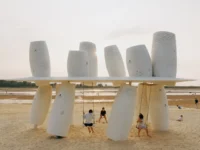 White perforated cylinders above and below a thin roof plane with two swings on a sandy beach at dusk, Seaside Swing Installation by XBTW OFFICE