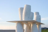Side view of the cylinders tilting at different angles above and below the flat roof against a blue sky, Seaside Swing Installation by XBTW OFFICE
