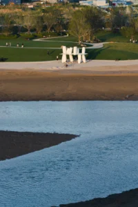 Shallow tidal water in the foreground with the installation on the far shore below the park, Seaside Swing Installation by XBTW OFFICE