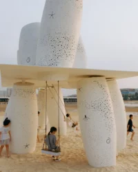 Children playing among the cylinders and on a swing hanging below the roof plane, Seaside Swing Installation by XBTW OFFICE