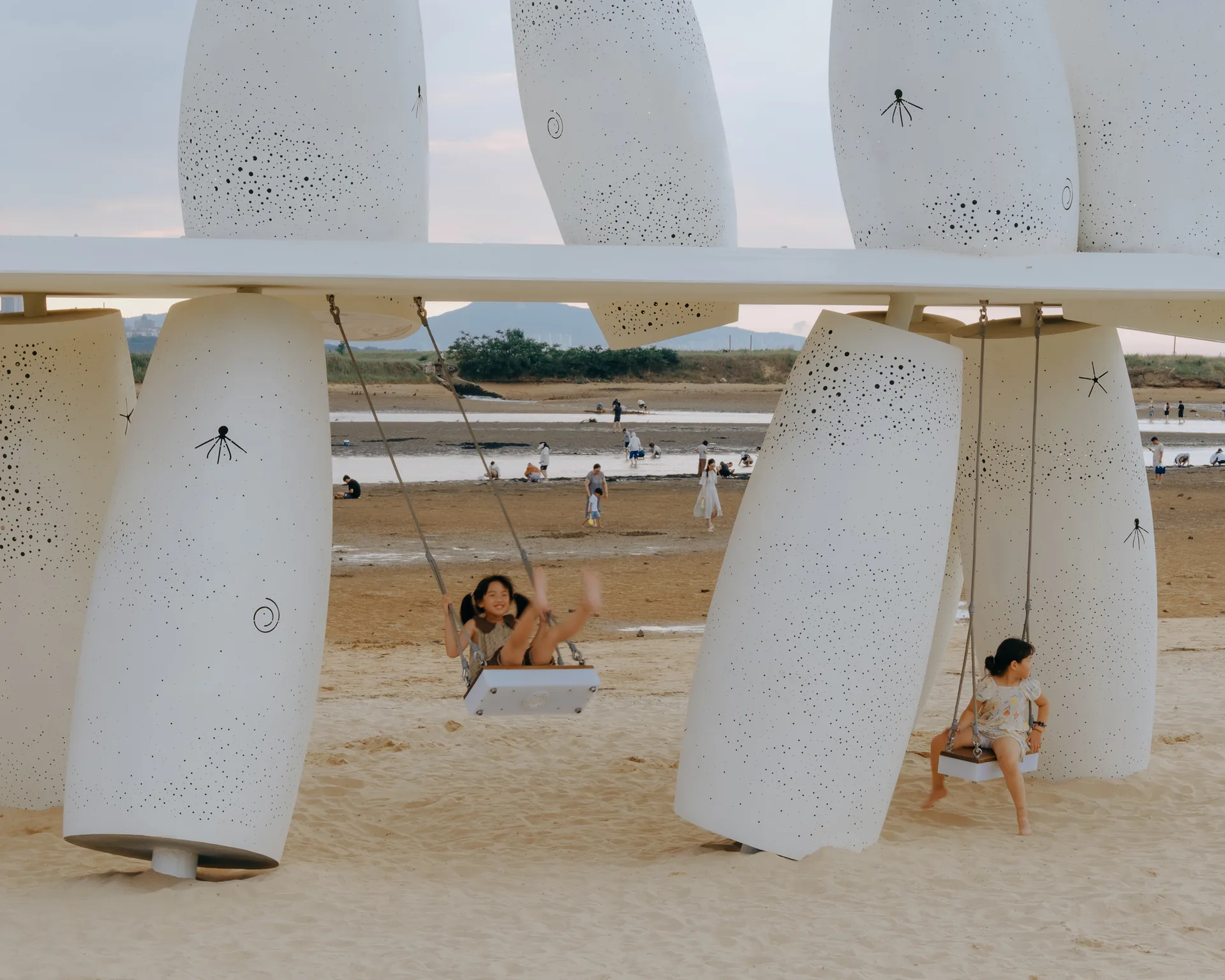 Two people on swings between leaning perforated columns with the beach behind, Seaside Swing Installation by XBTW OFFICE