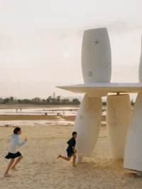 Children running across the sand past the base of the cylinders in evening light, Seaside Swing Installation by XBTW OFFICE