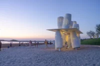 Installation glowing from within at blue hour with people walking on the beach, Seaside Swing Installation by XBTW OFFICE